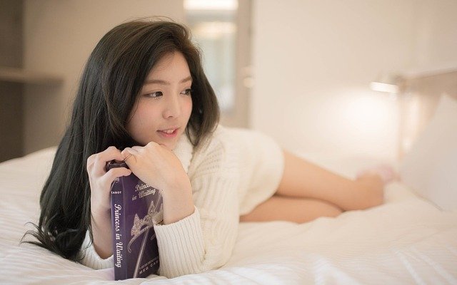 Žena v bielom pletenom svetri leží na posteli a v rukách drží knihu.jpg